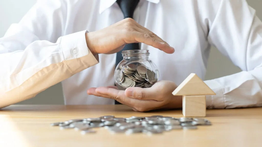 Un homme en chemise blanche protégeant de ses mains un bocal en verre rempli de pièces de monnaie, posé à côté d'une petite maison en bois, symbolisant la sécurité des enveloppes d'épargne.