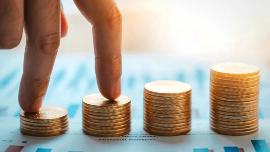 Deux doigts mimant une marche en grimpant sur des piles croissantes de pièces de monnaie dorées, illustrant la régularité de l'investissement mensuel et les petits pas vers la richesse.